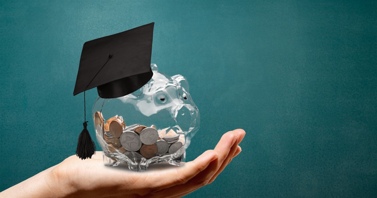 Partially full glass piggy bank with graduation cap, being held in an outstretched hand