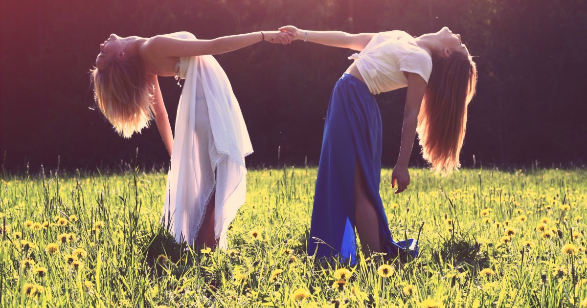 Two young girls leaning all the way back while holding hands in a field with sunlight